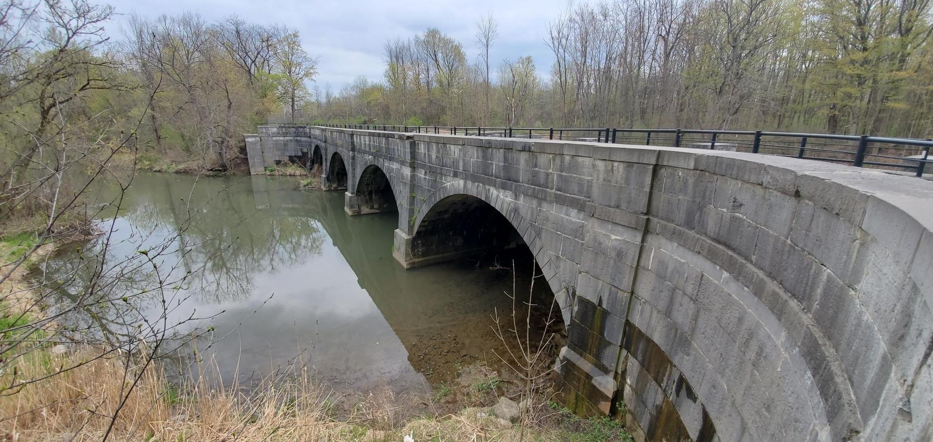 A stone arch bridge spans a calm river, surrounded by trees on an overcast day.