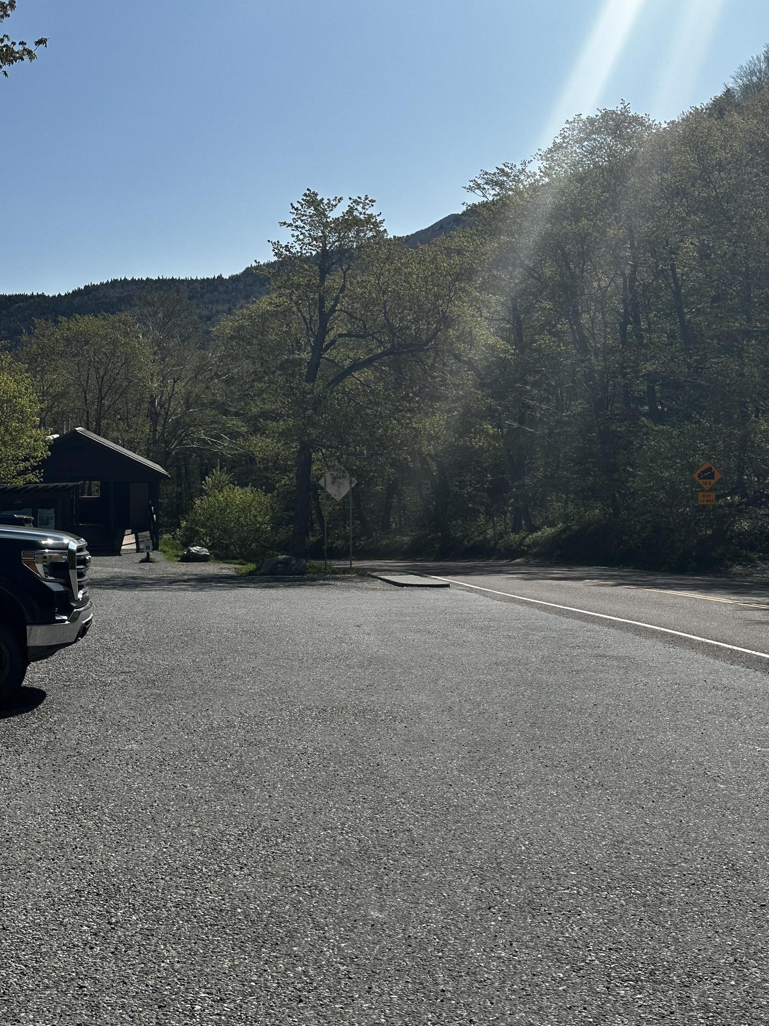 Sunny parking lot with a vehicle, building, and wooded mountainside.