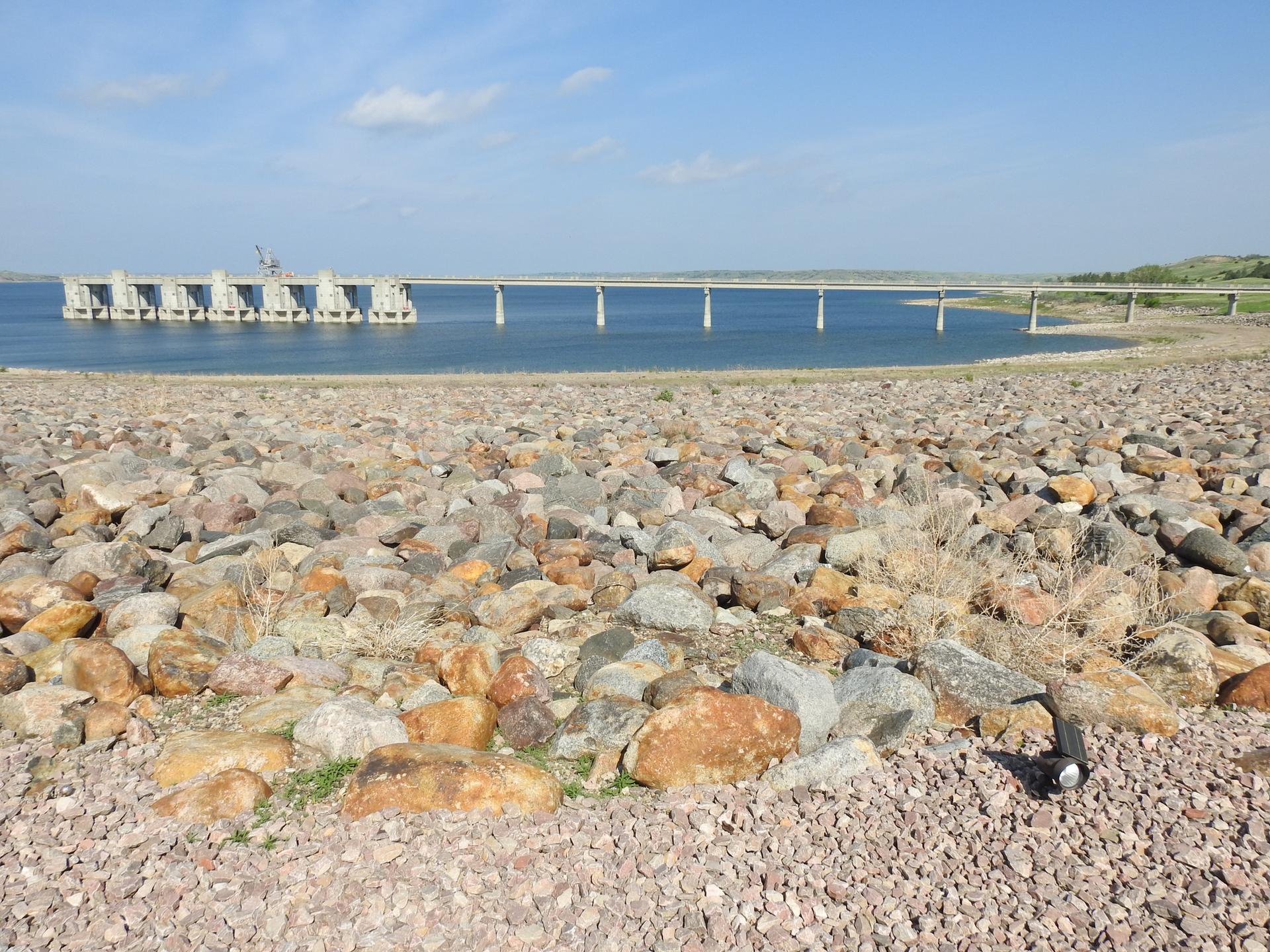Rocky shore in foreground, wide lake with distant dam and bridge under clear sky.