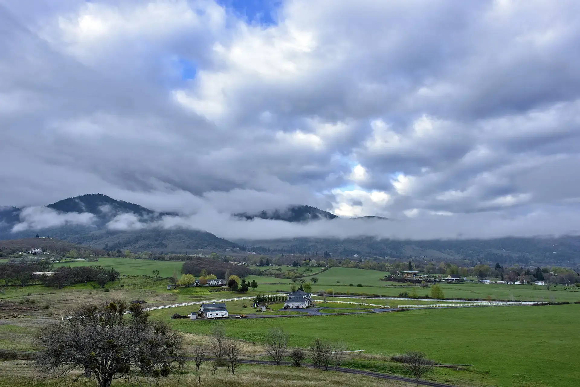 Green fields and scattered houses under a cloudy sky with misty mountains.