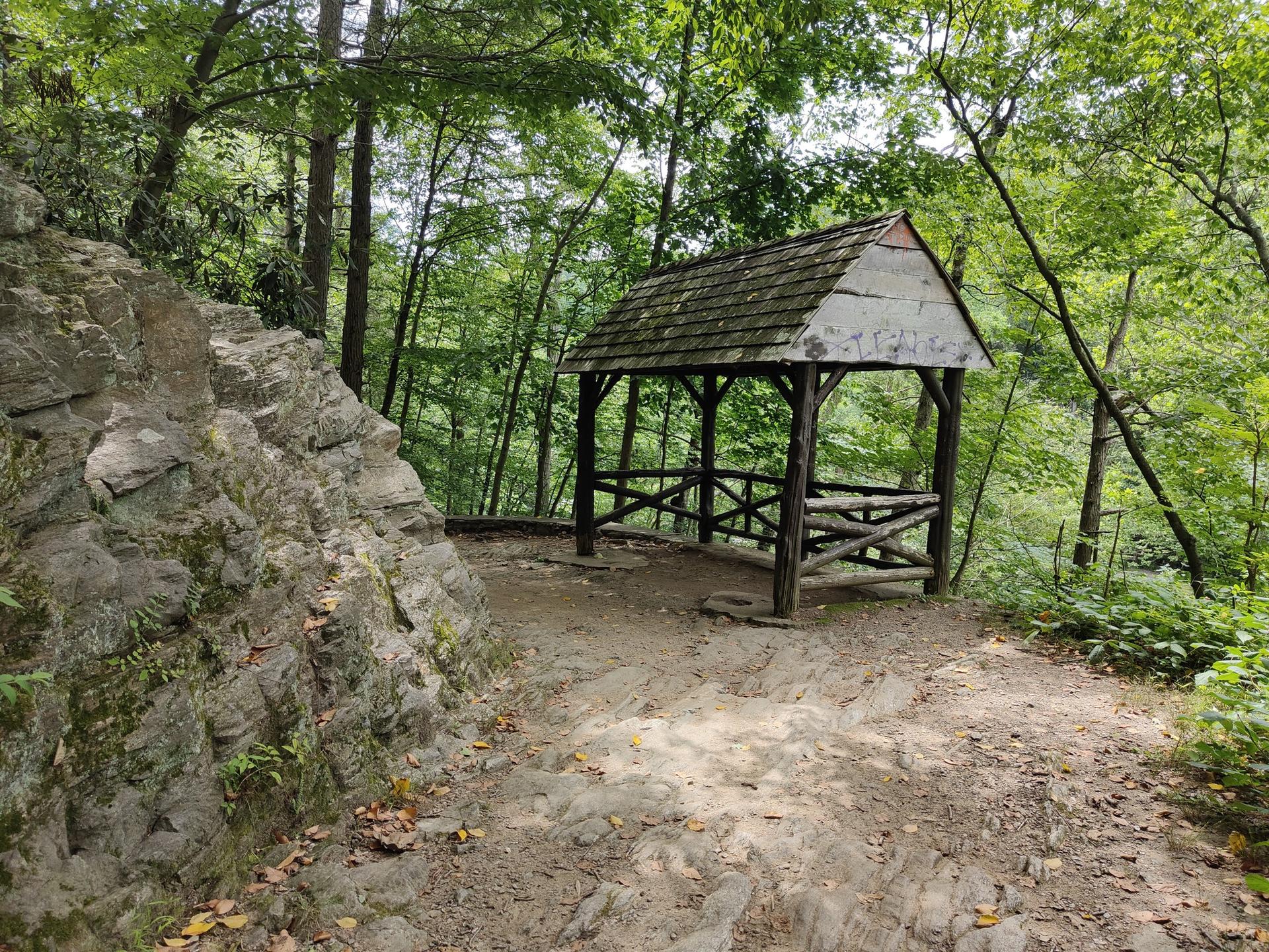 A rustic wooden gazebo sits beside a rocky outcrop in a green forest.