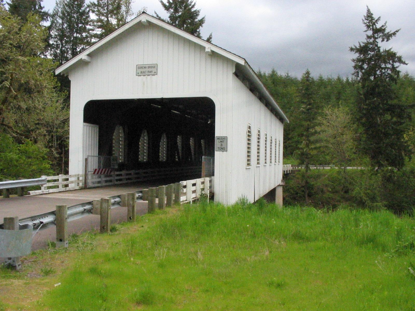 White covered bridge with green grass and dark forest under a cloudy sky.