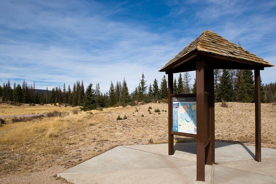 Wooden information kiosk in a sunny, open field with pine trees.