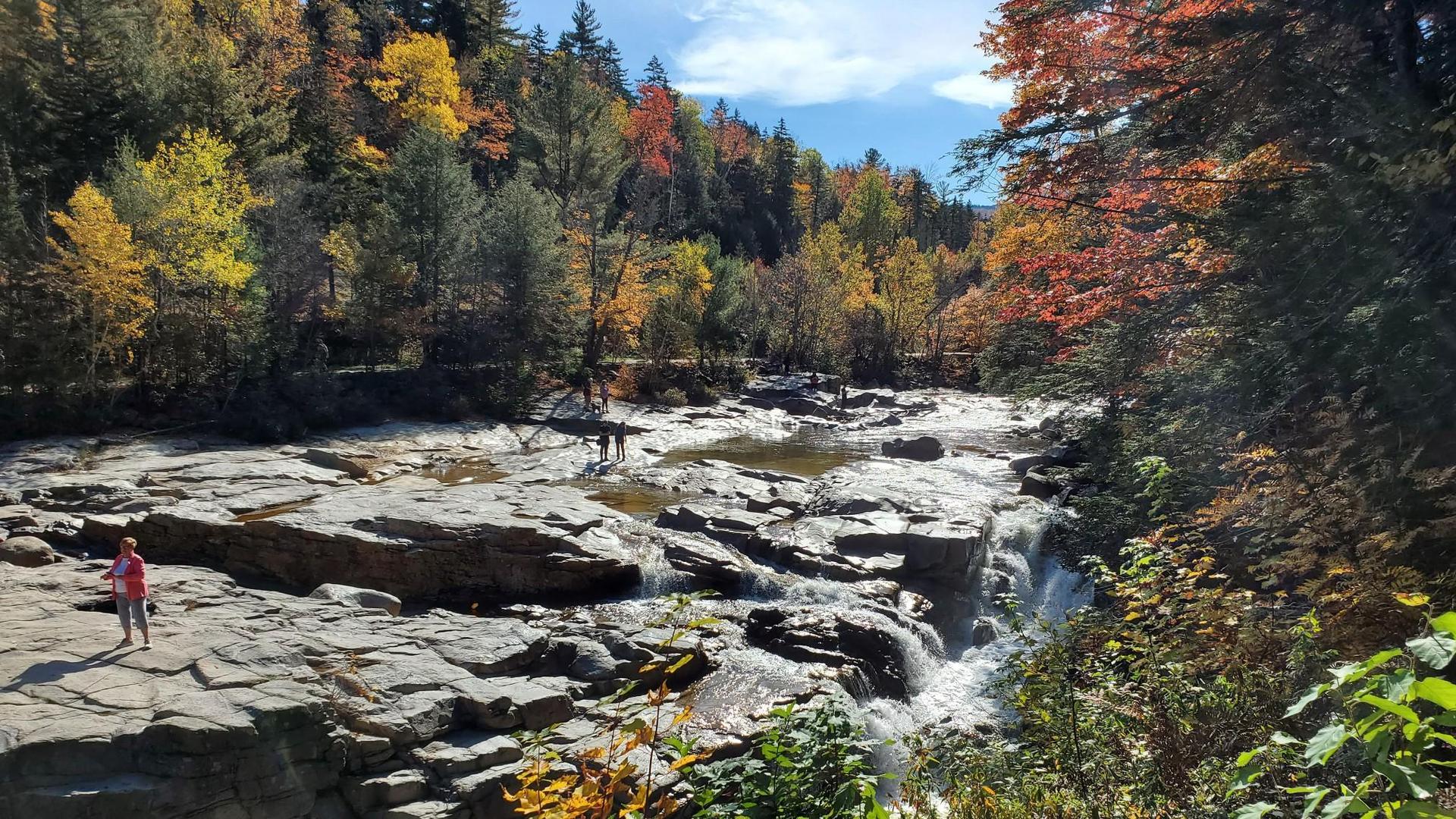 Sunny river with small waterfalls, people on rocks, vibrant autumn forest.