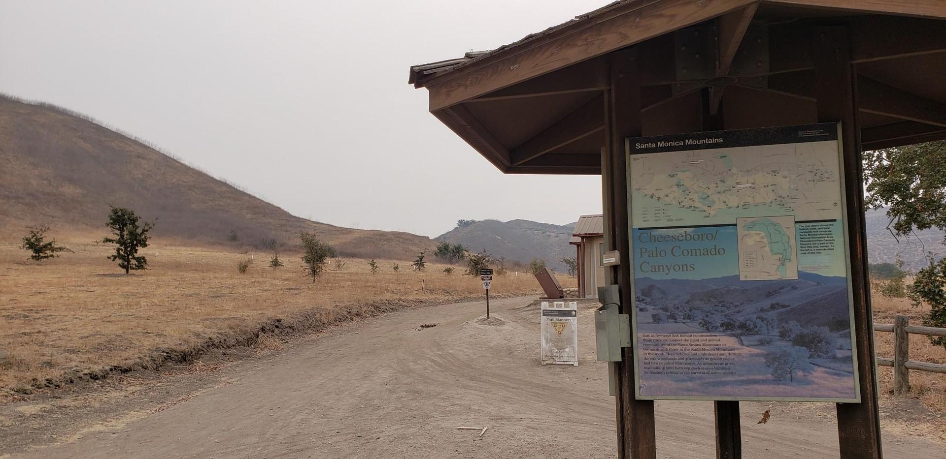 Information kiosk next to a dirt road, leading to dry hills under an overcast sky.