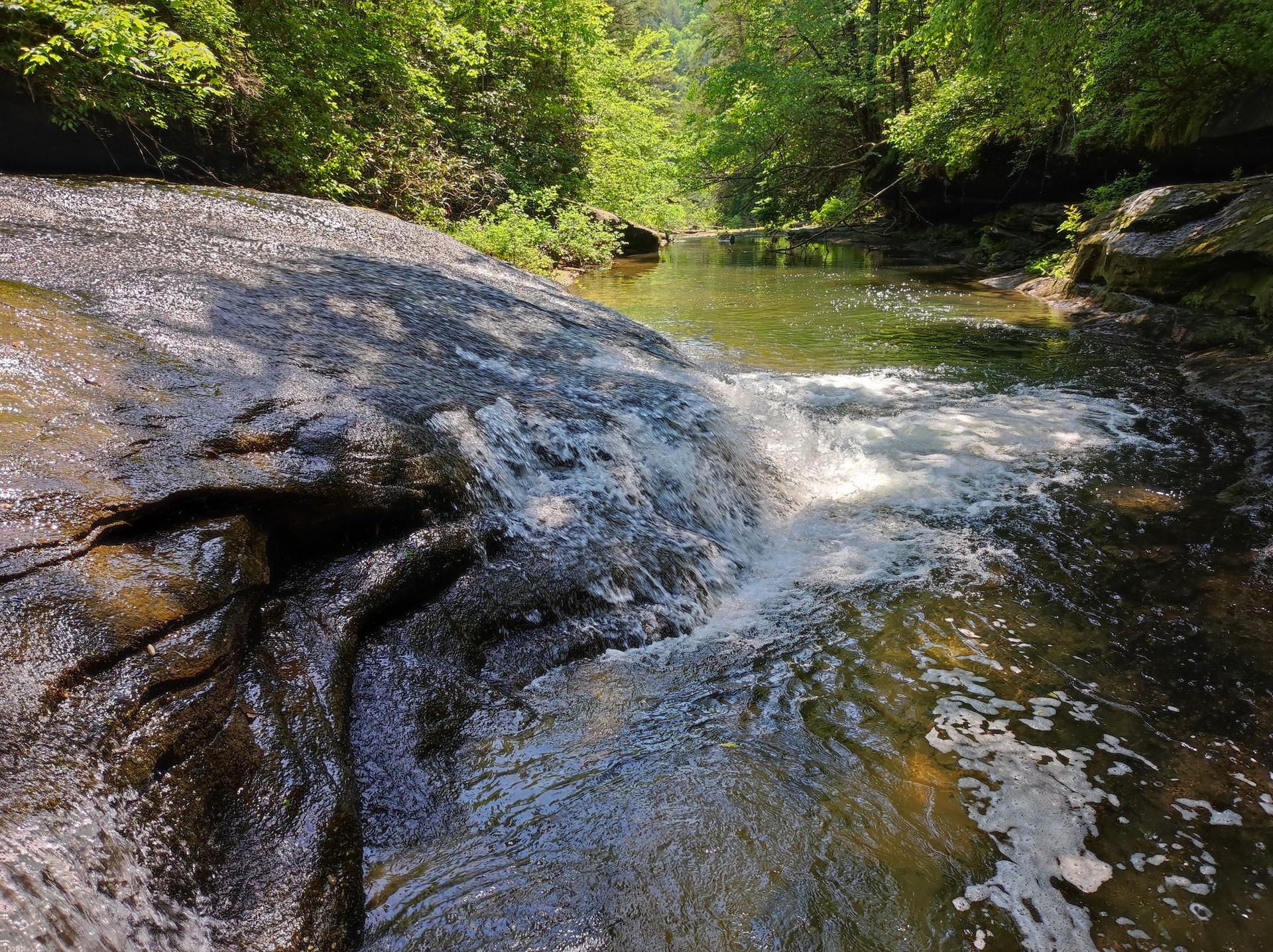 Small waterfall cascading over dark rocks into a clear, sunlit stream.