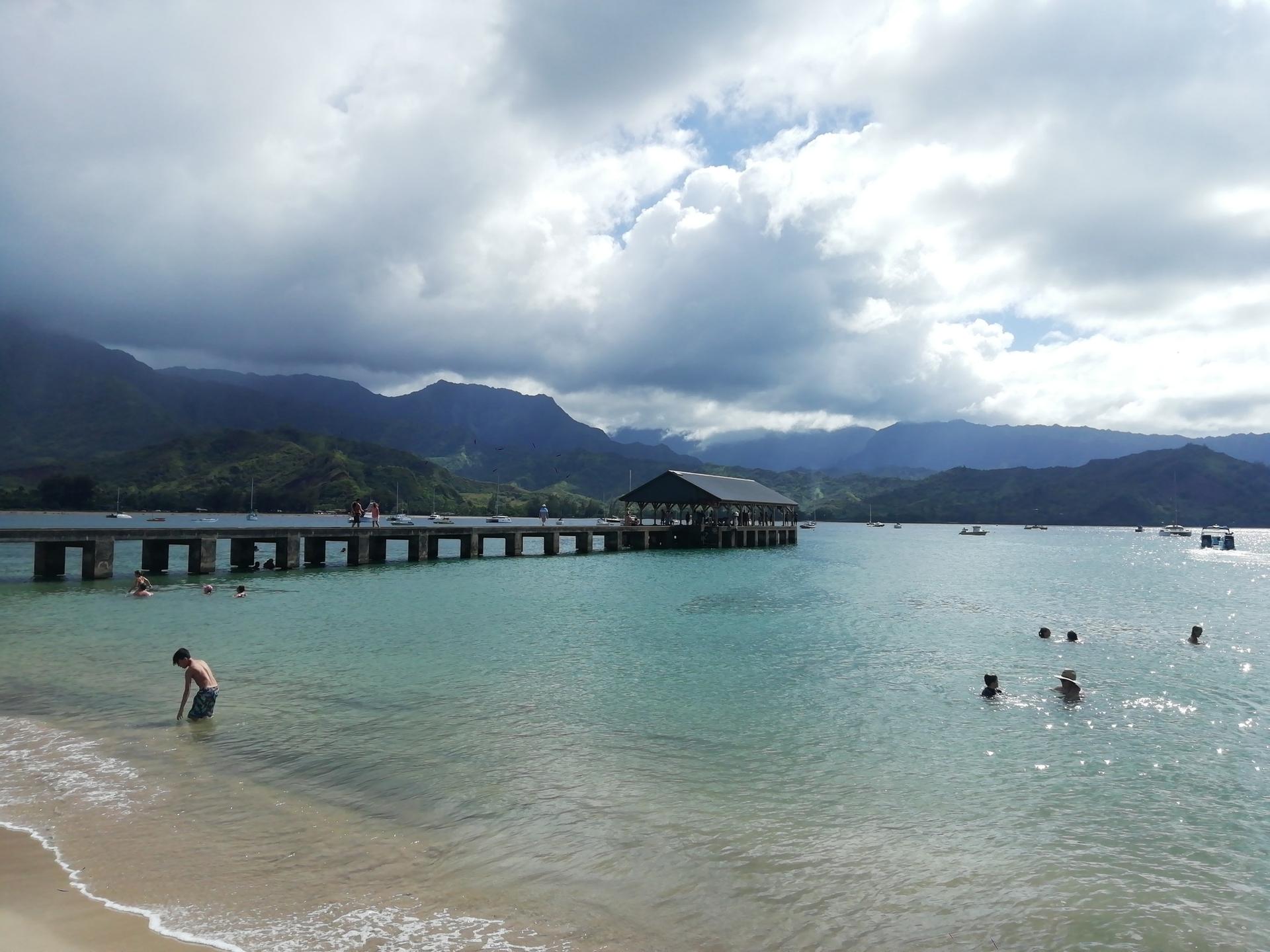 People swim near a pier and sandy beach, with mountains under a cloudy sky.