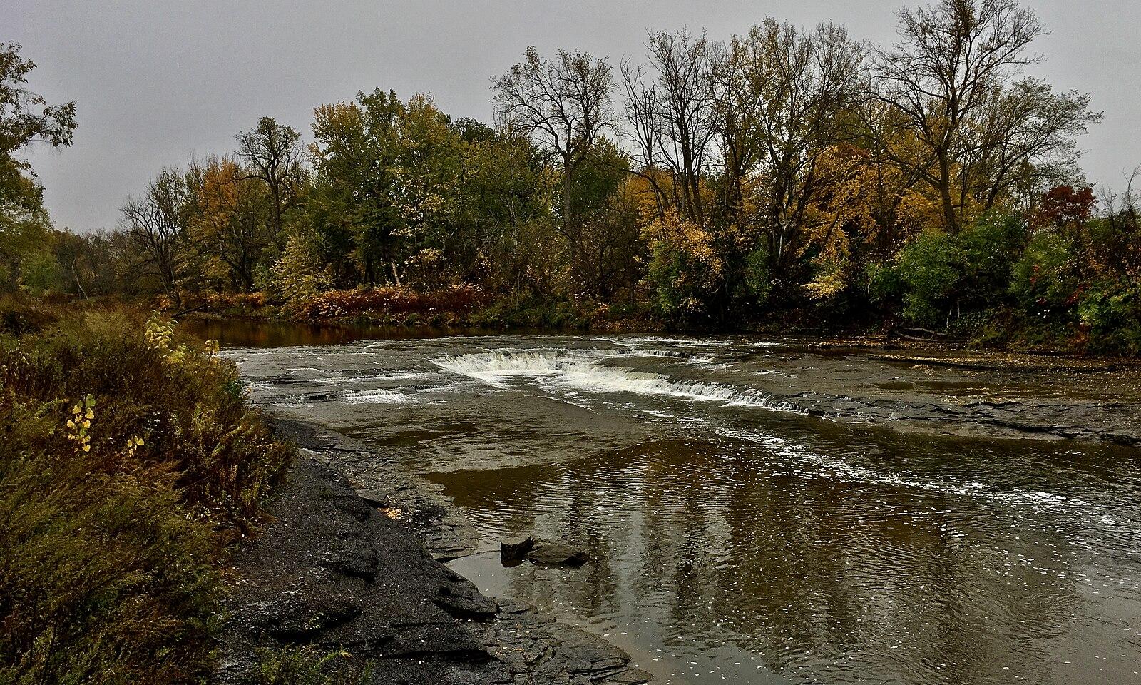 A wide river with small rapids flowing past autumn trees.