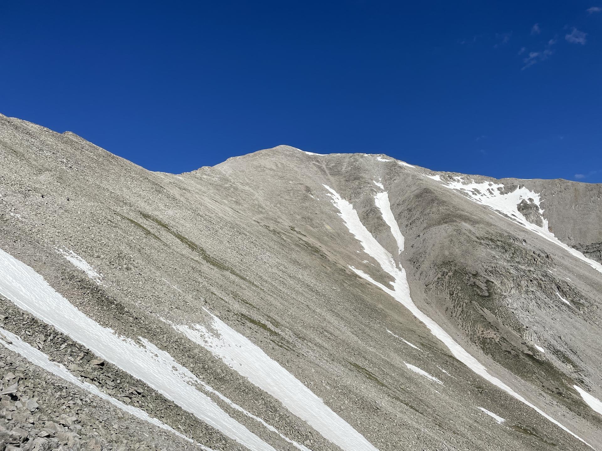 Steep, rocky mountain slope with snow patches under a bright blue sky.