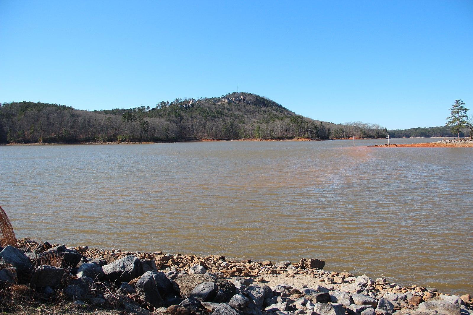 Wide lake with rocky foreground and distant forested hills under clear sky.