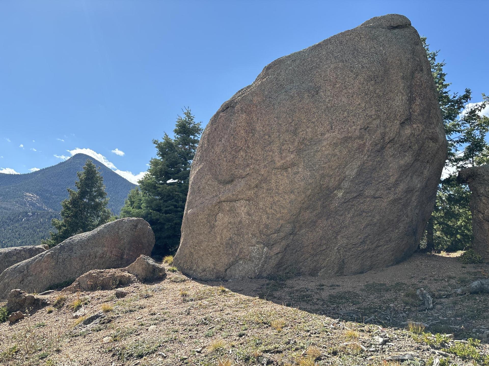 Large boulder on a sunny day with mountains and pine trees.