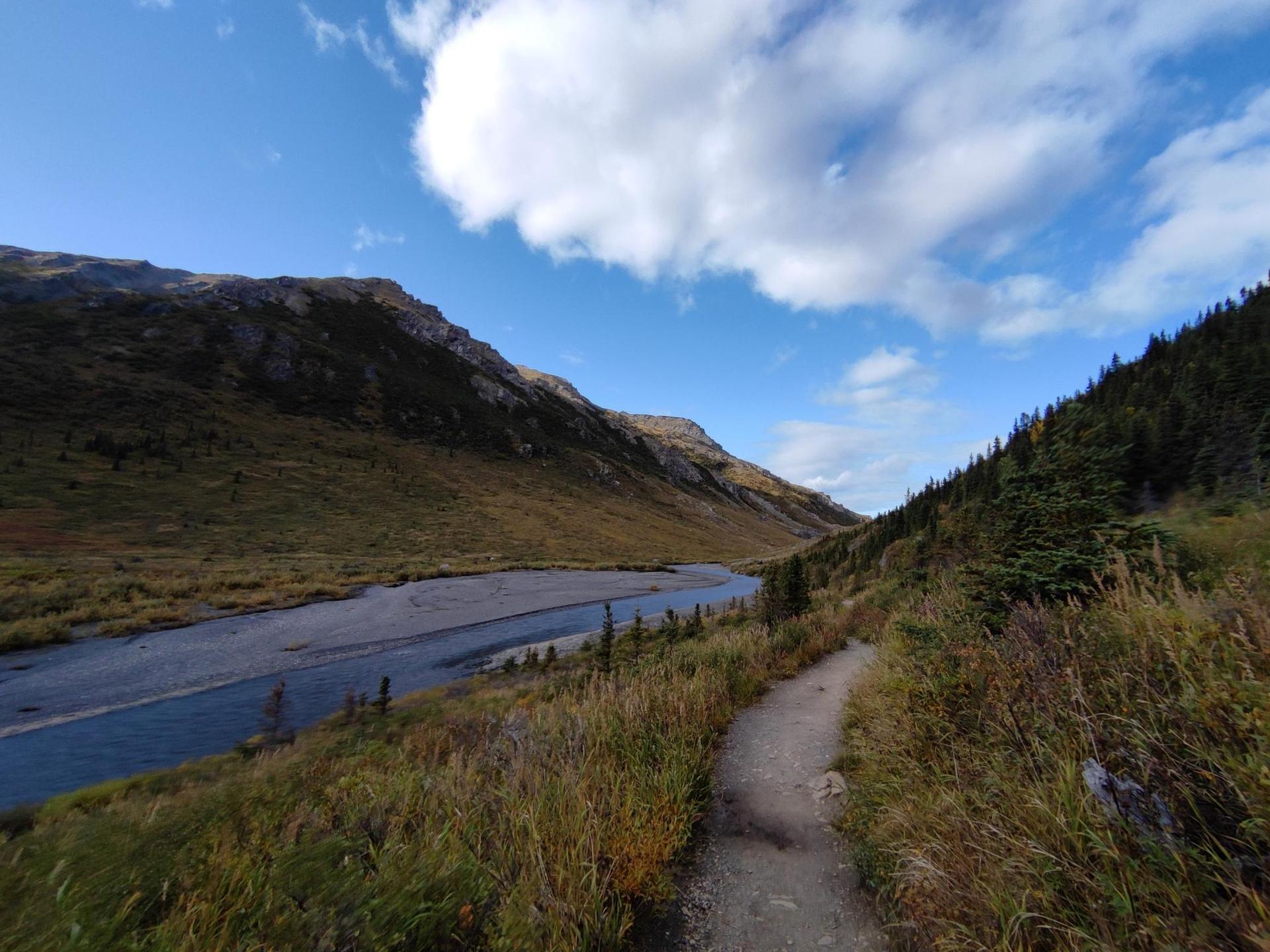 Dirt path beside a river in a green valley under a blue sky.