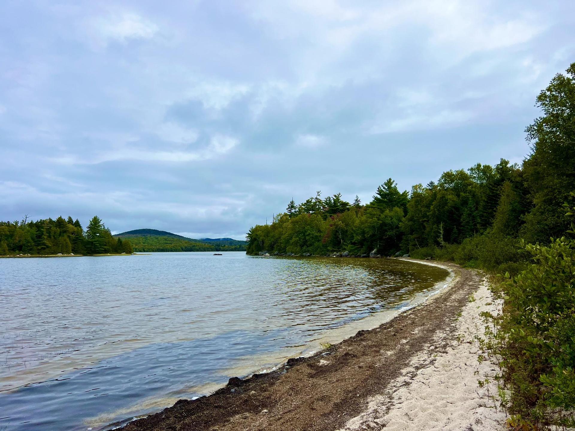 Wide lake with a sandy and rocky shoreline under a cloudy sky.