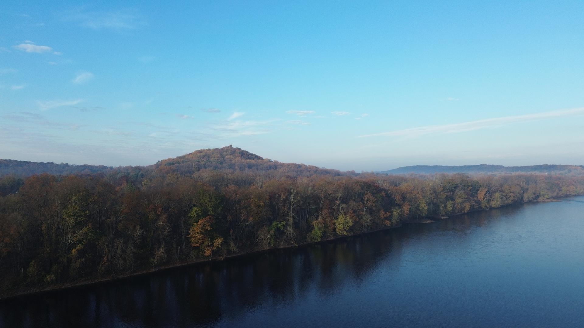 An aerial view of a wide river next to forested hills under a blue sky.