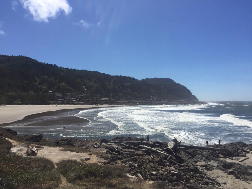 Sunny beach with crashing waves, forested hills, and distant people.