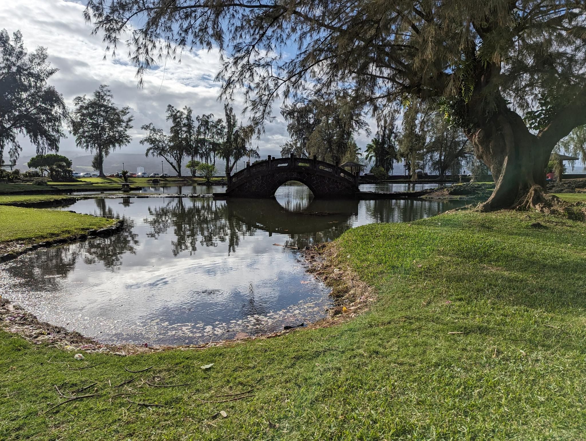 Stone arch bridge over a reflective pond, surrounded by lush green trees. - Liliuokalani Gardens – Rainbow Falls loop from Wailoa River State Recreation Area