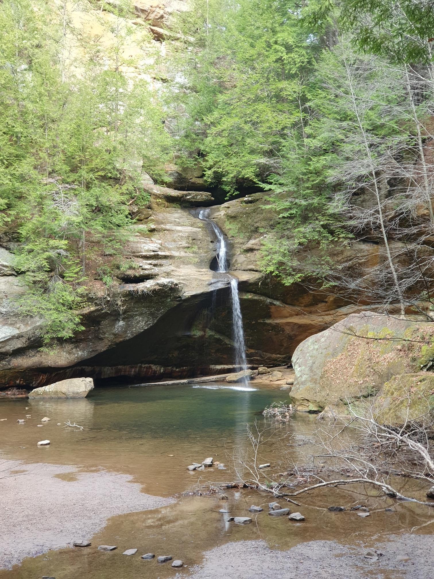 Tall waterfall plunges into a clear pool, surrounded by rocky cliffs and green trees.