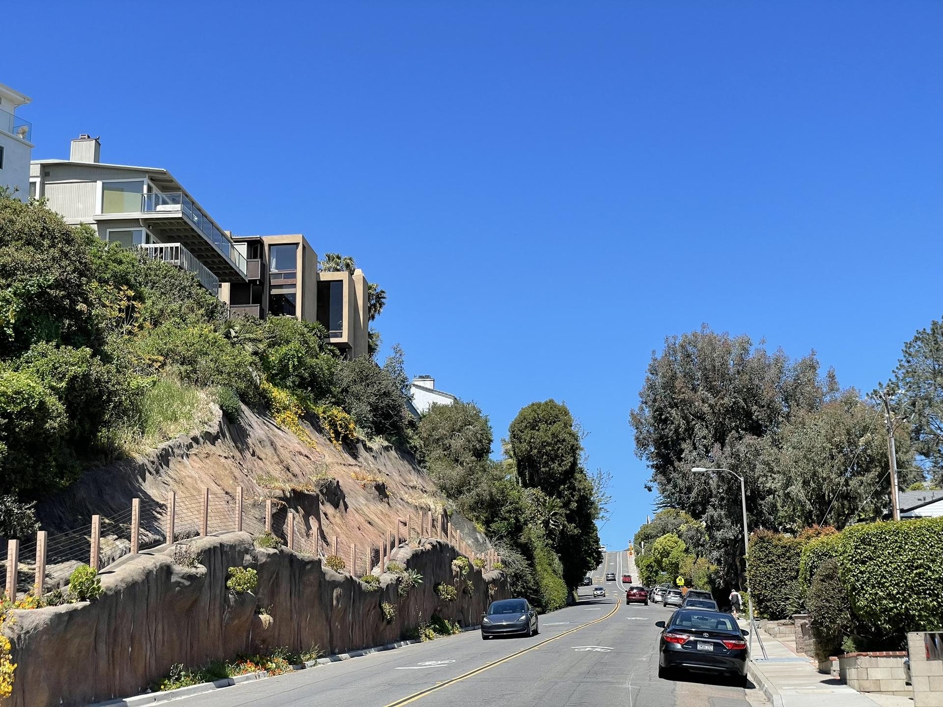 Cars on a road next to a steep, green hillside with modern houses.