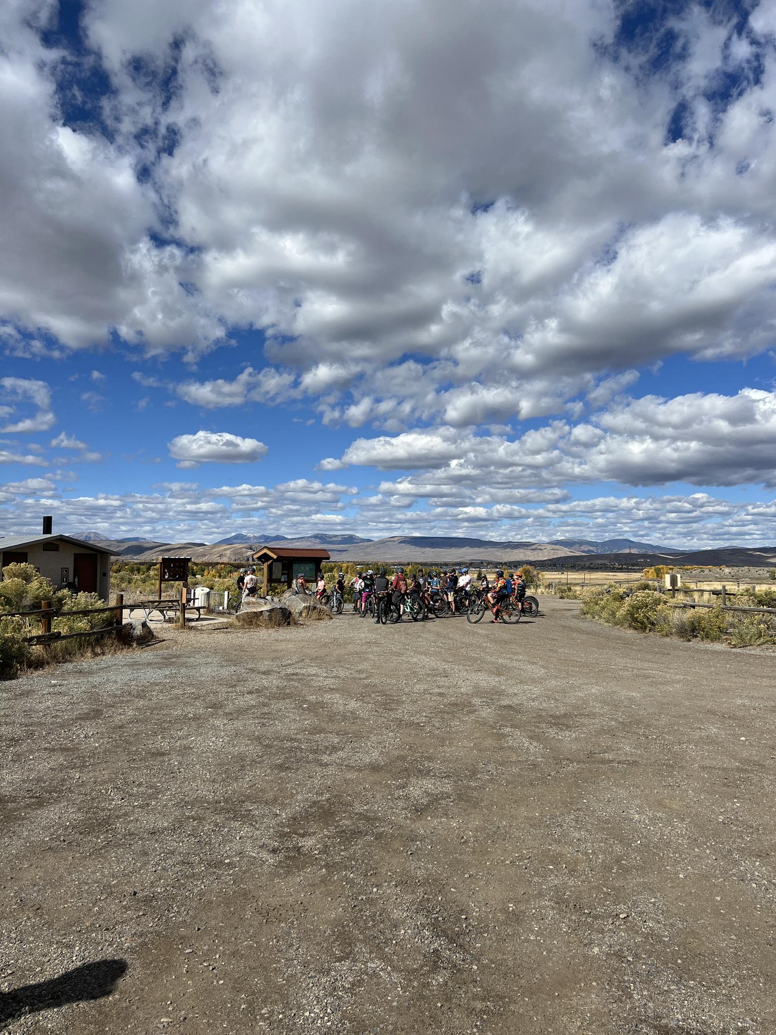 A group of cyclists gathers on a dirt road under a cloudy blue sky.