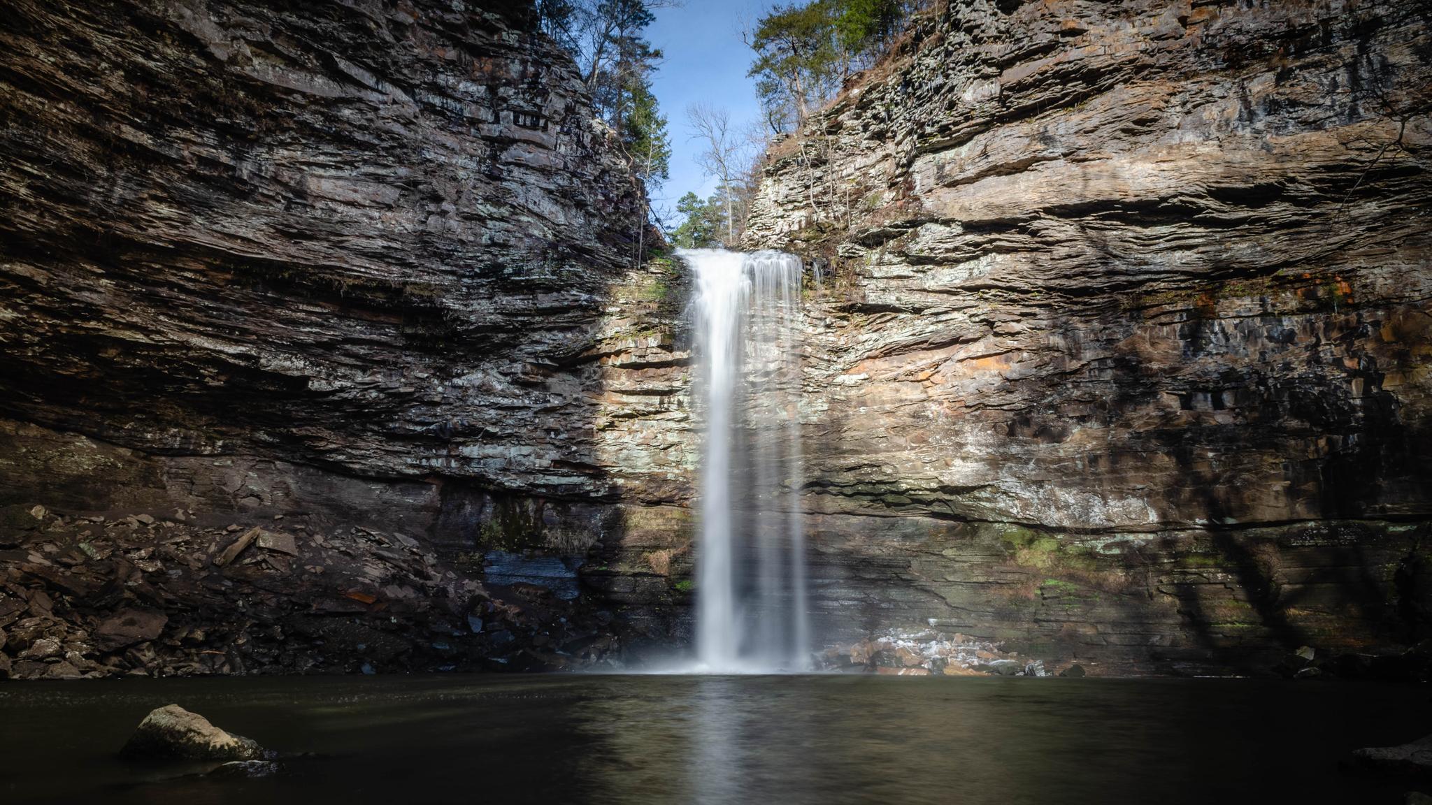 Tall waterfall plunges into a dark pool surrounded by rocky cliffs and trees.
