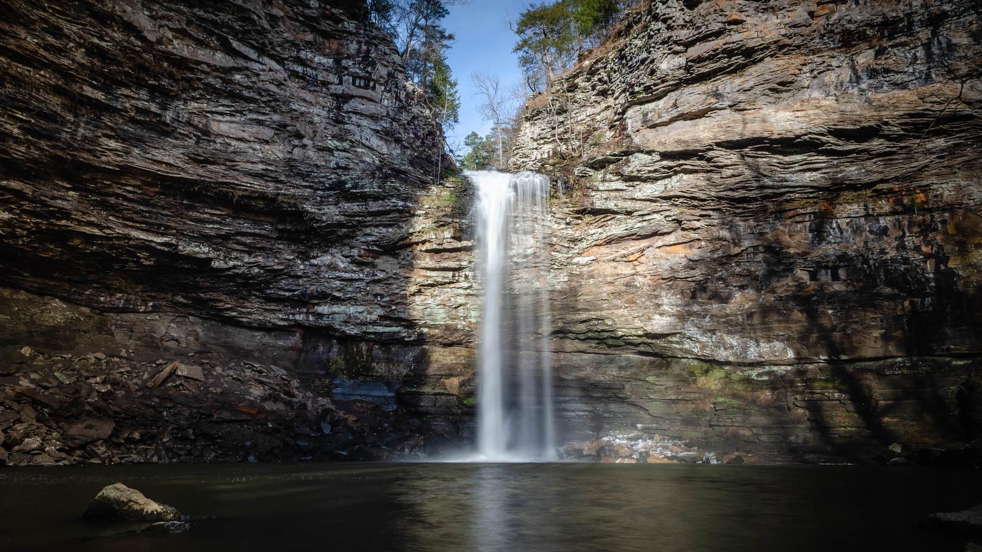 Tall waterfall plunges into a dark pool surrounded by rocky cliffs and trees.