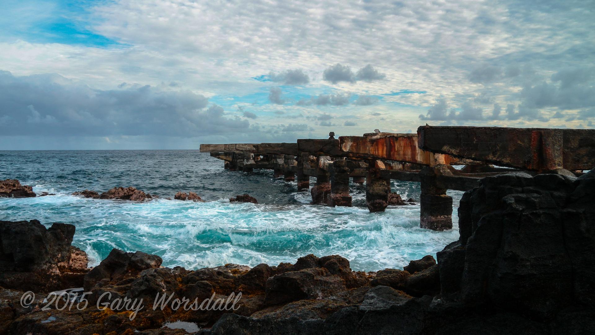 A weathered concrete pier extends into the turbulent ocean under an overcast sky.