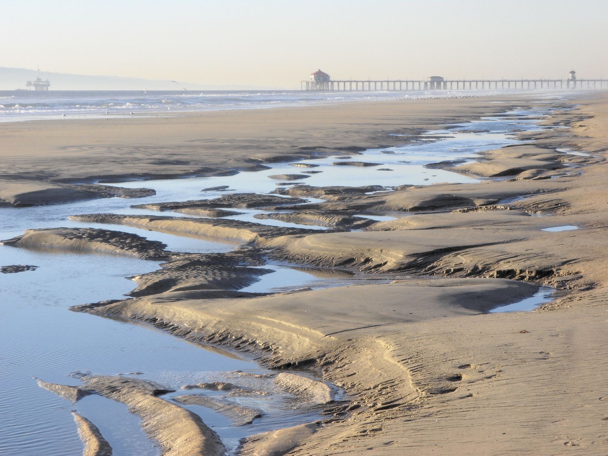 Sandy beach with tidal pools and a distant pier under hazy light. - Huntington Beach Bike Trail