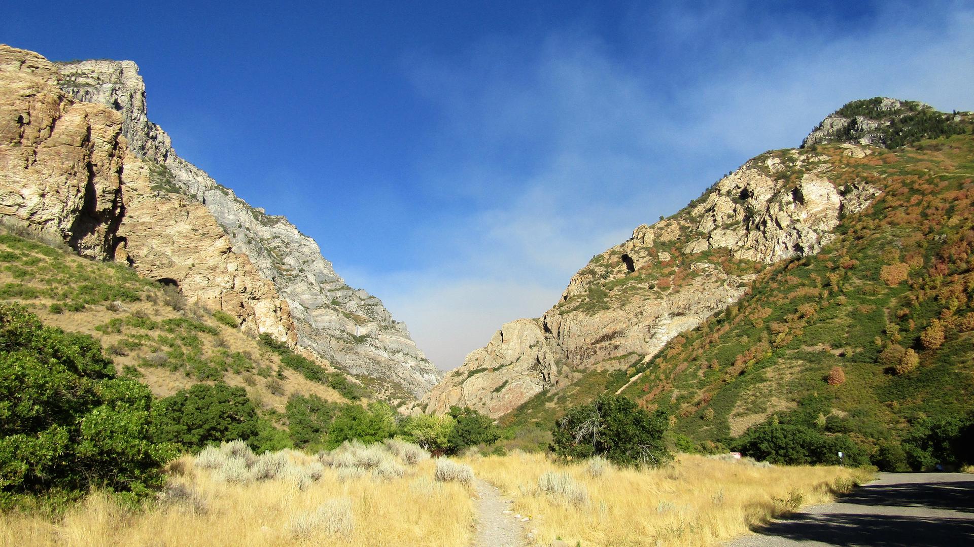 Wide canyon with steep rocky slopes and a dirt path under a blue sky.