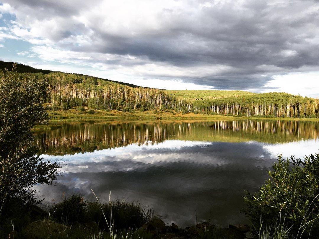 Serene lake reflecting forested hills and dramatic cloudy sky with patches of light.