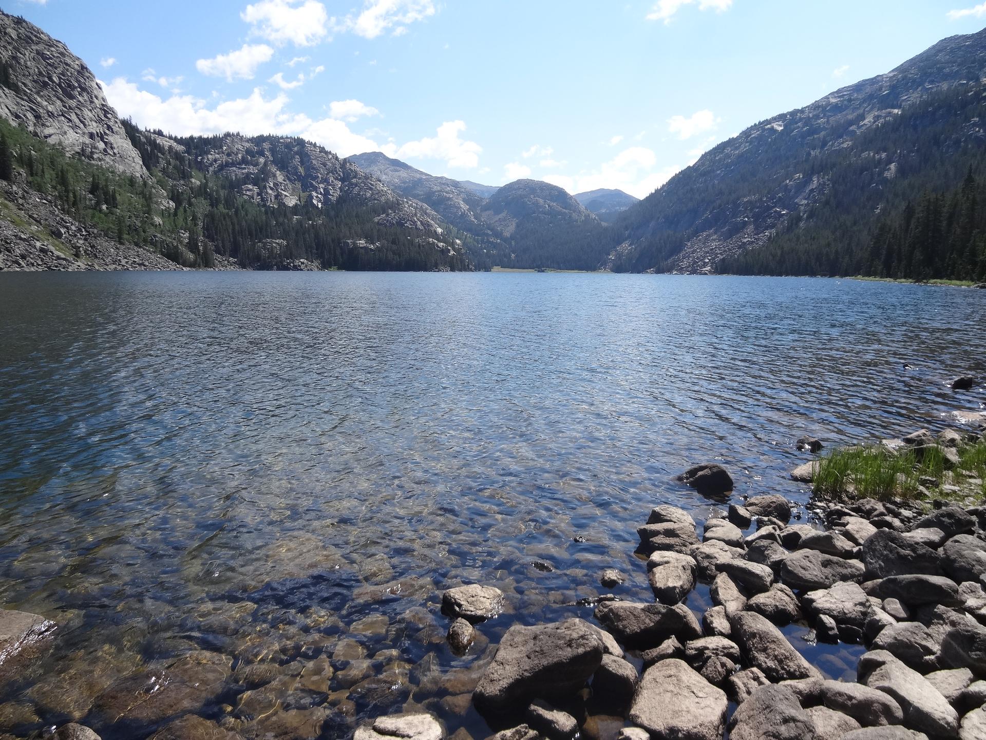 Clear mountain lake with rocky shore and distant mountains under blue sky.