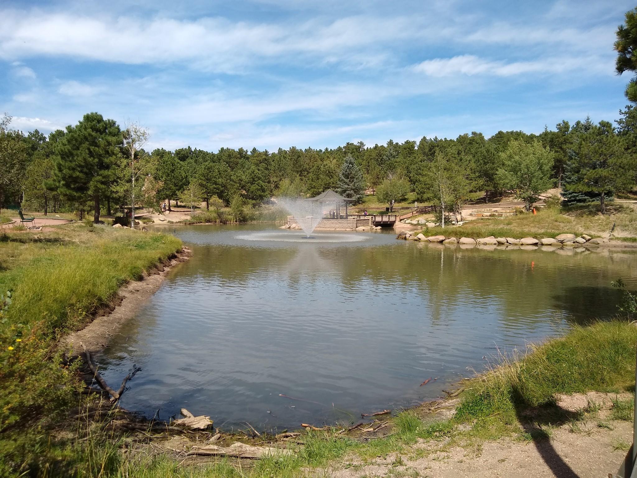Lake with a fountain, surrounded by green trees under a blue sky.
