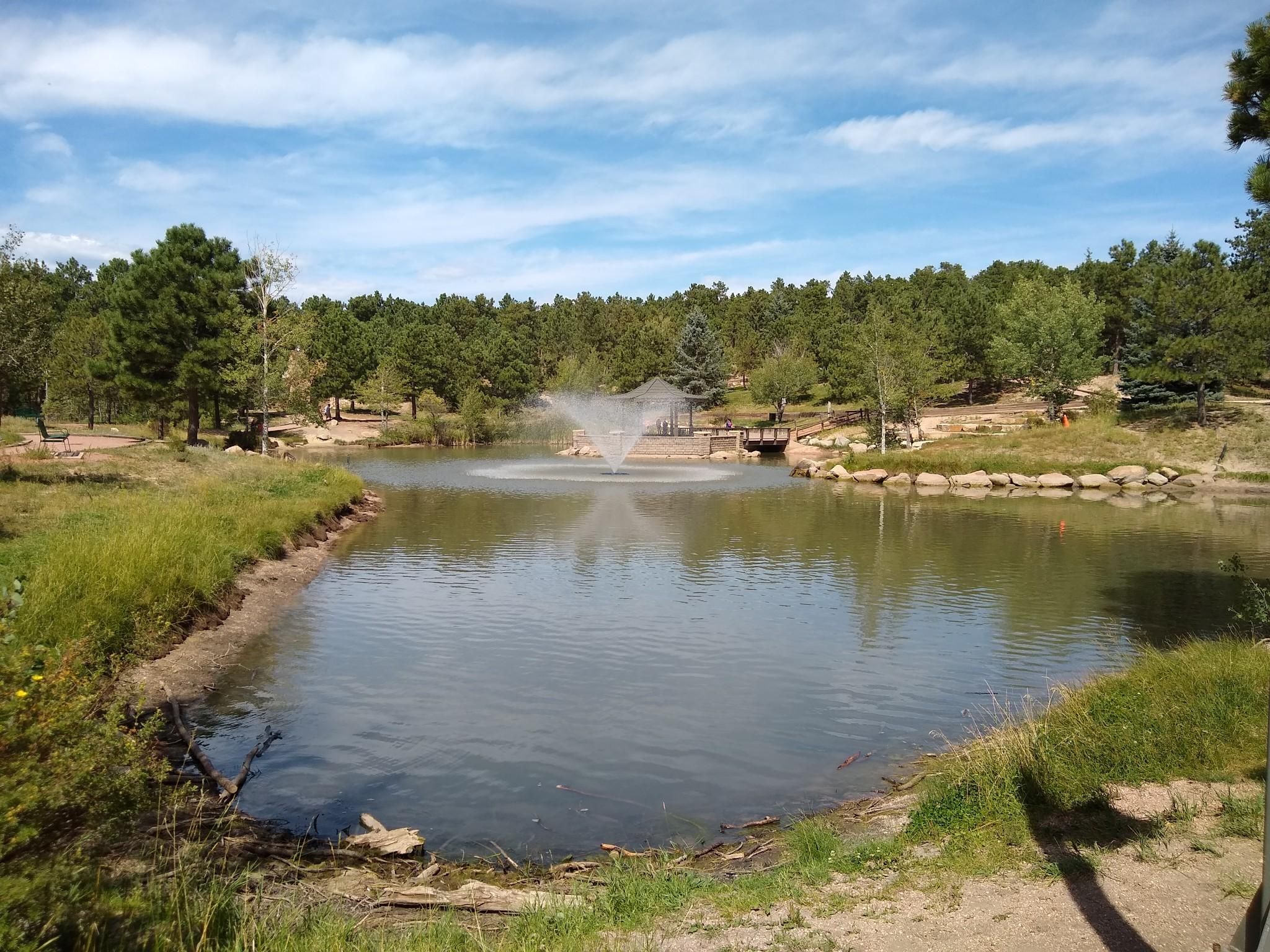 Lake with a fountain, surrounded by green trees under a blue sky. - Fox Run Loop Trail