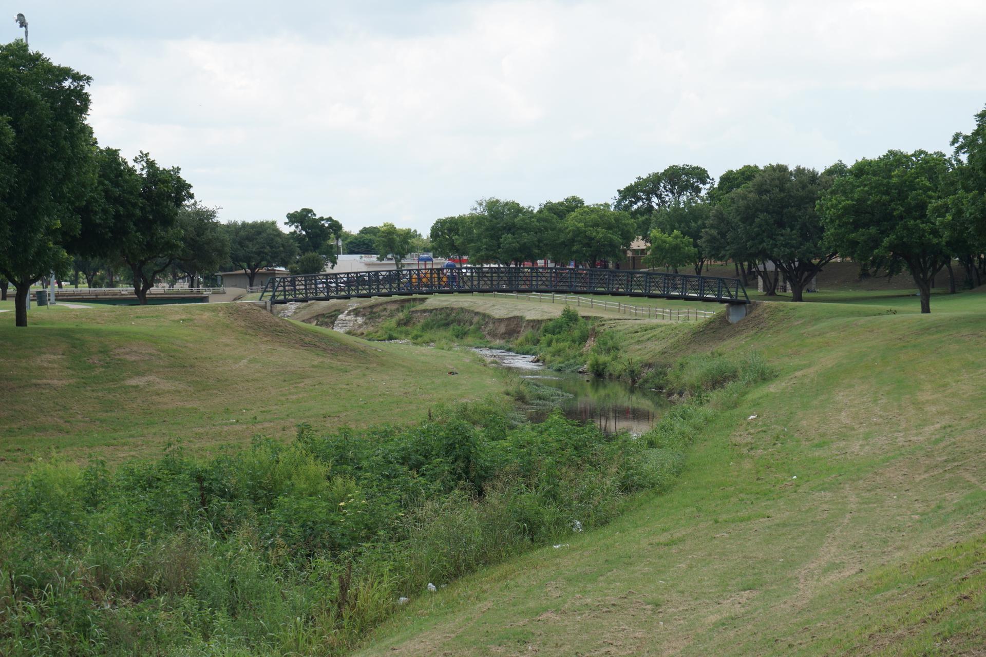 Pedestrian bridge over a creek in a green park with people walking.