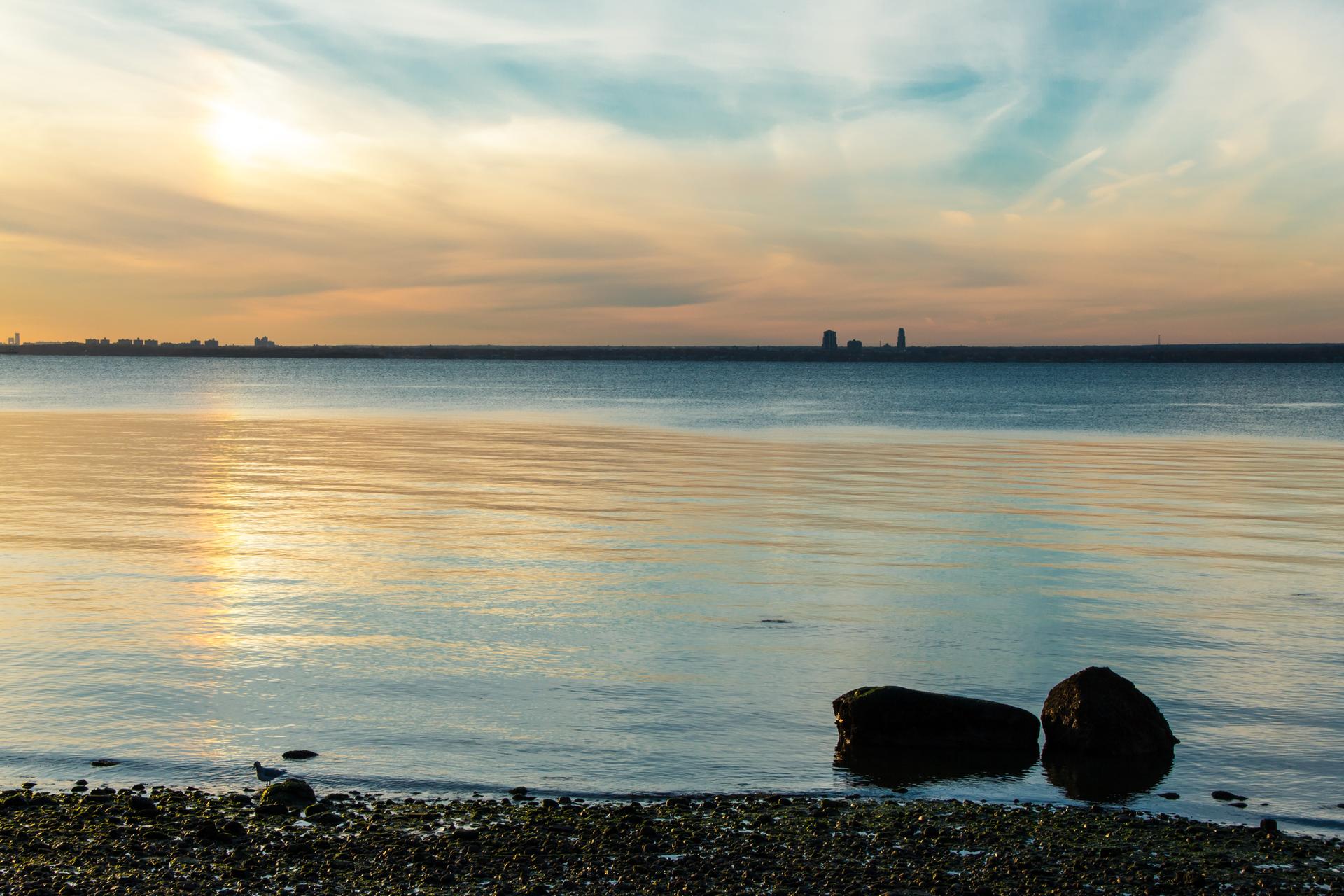Golden sunset light reflects on calm water with distant city and rocky shore.