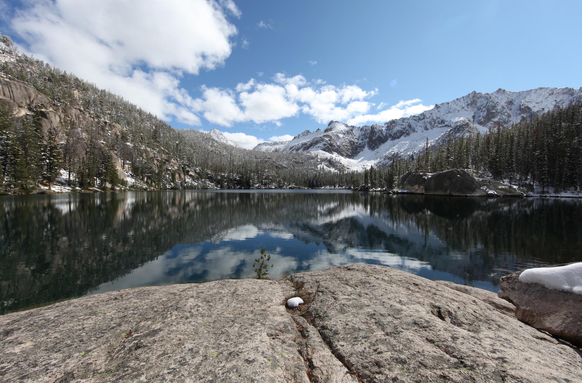 Clear mountain lake reflecting snowy peaks and blue sky, with a rock in foreground.