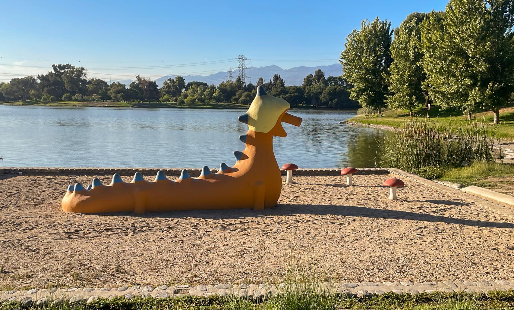 Orange dragon playground structure on sand next to a lake under bright sun.