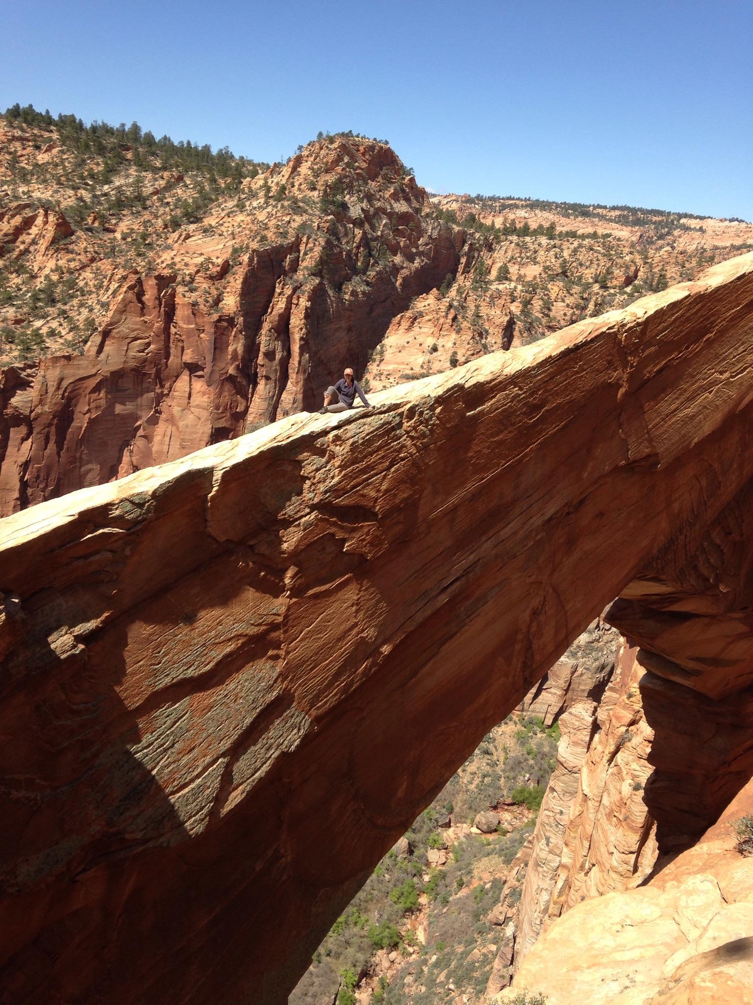 A person sits on a natural rock arch above a canyon, bright daylight.