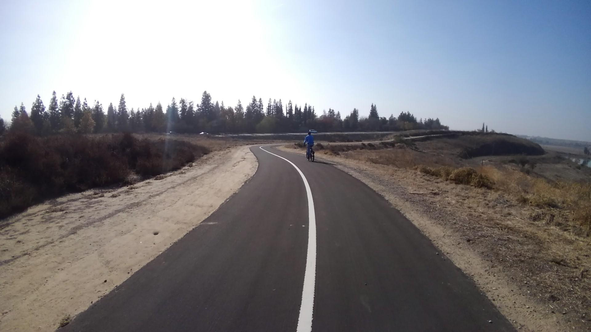 A cyclist rides on a paved path next to dry hills on a sunny day.
