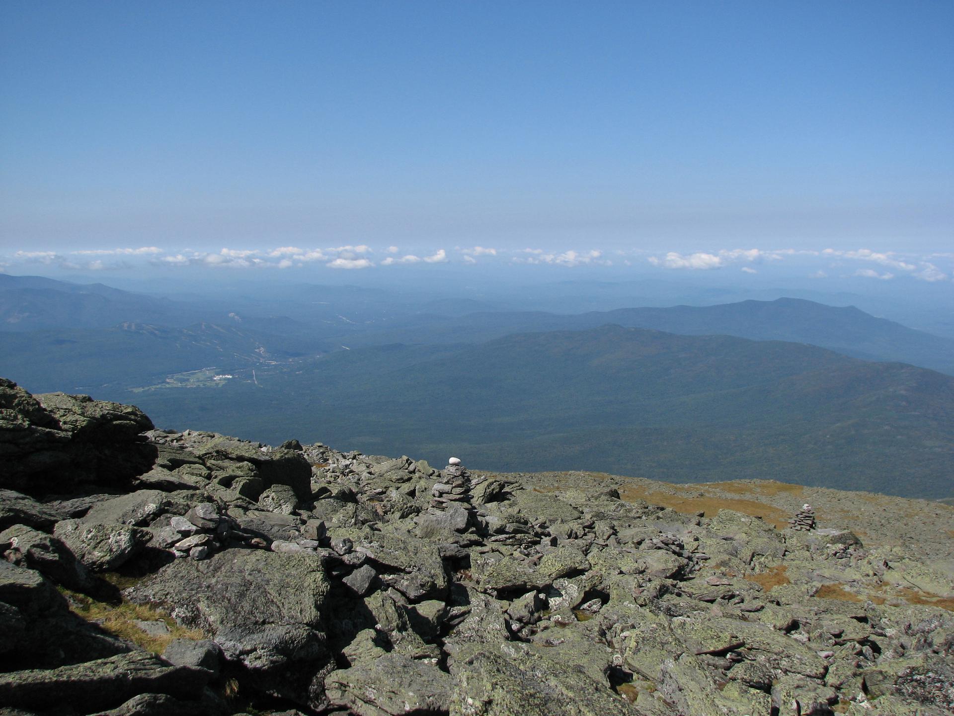 Sunny mountain summit with rocky foreground, distant peaks, and clear sky.
