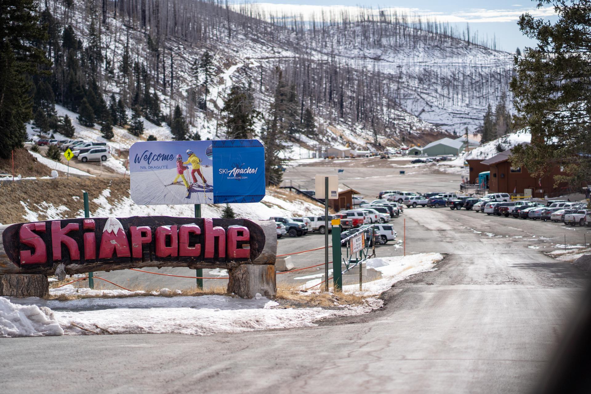 A large wooden sign and billboard at a snowy mountain resort entrance with parked cars.