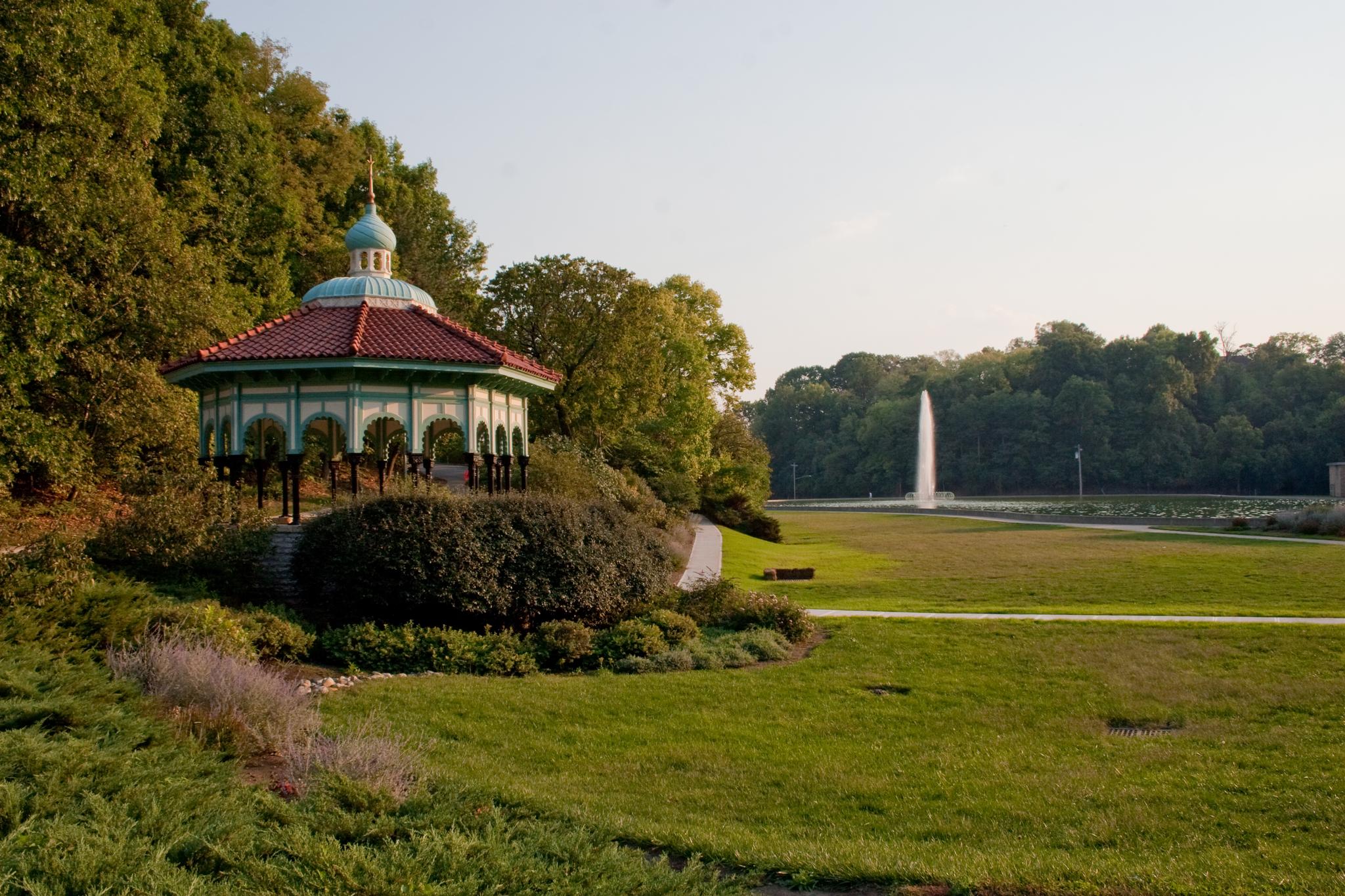 Ornate gazebo overlooks a park with a tall fountain in golden light.