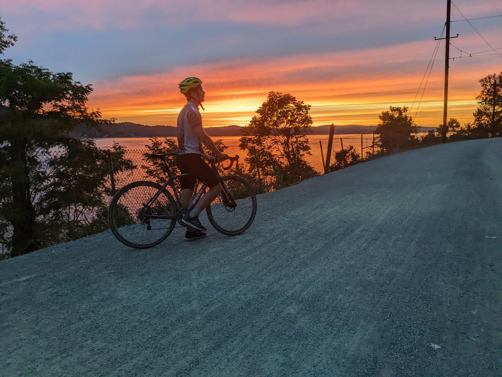 Cyclist on a road overlooking a lake at sunset, with orange and pink sky.