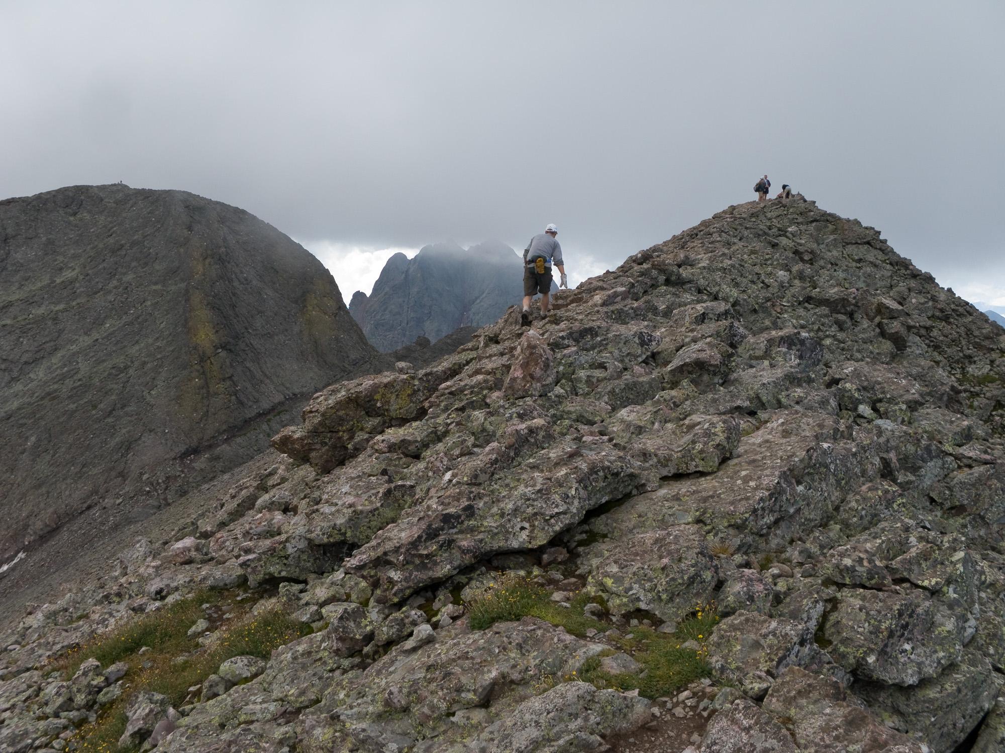 Hiker ascending a rugged, rocky mountain ridge under an overcast sky.