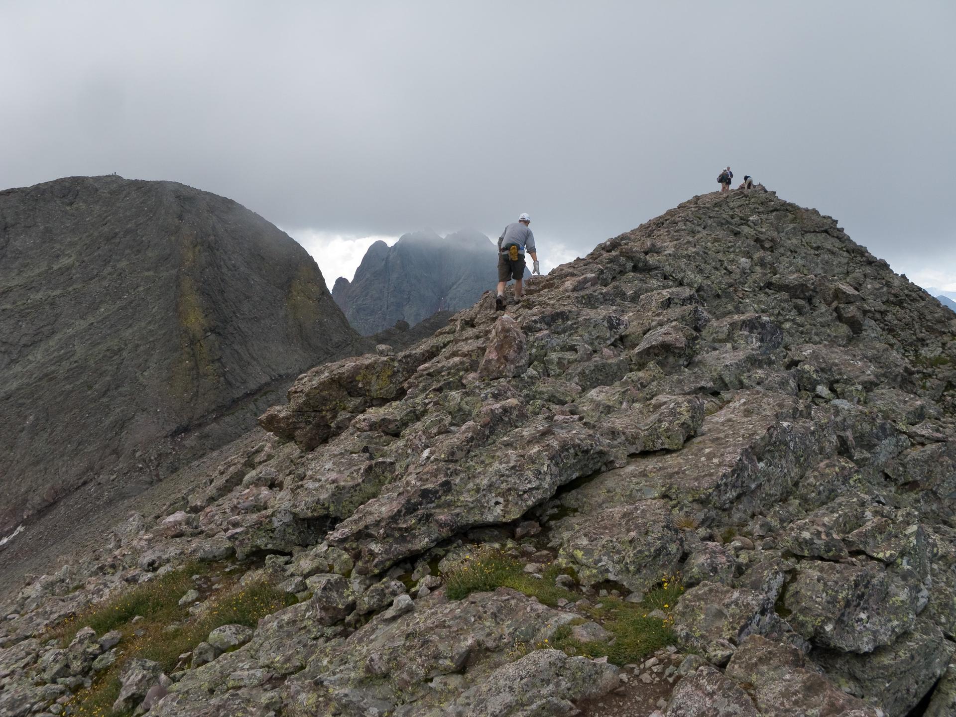 Hiker ascending a rugged, rocky mountain ridge under an overcast sky.