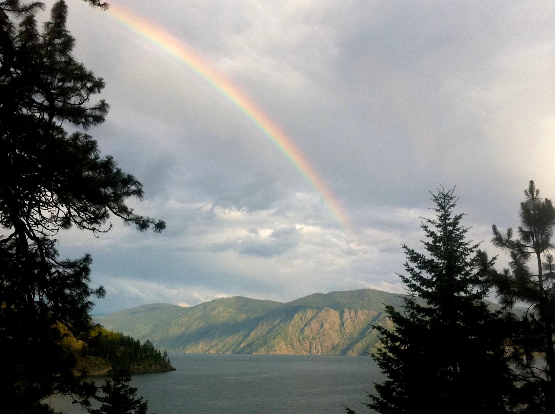 A vibrant rainbow arches over a serene lake and distant mountains under cloudy skies.