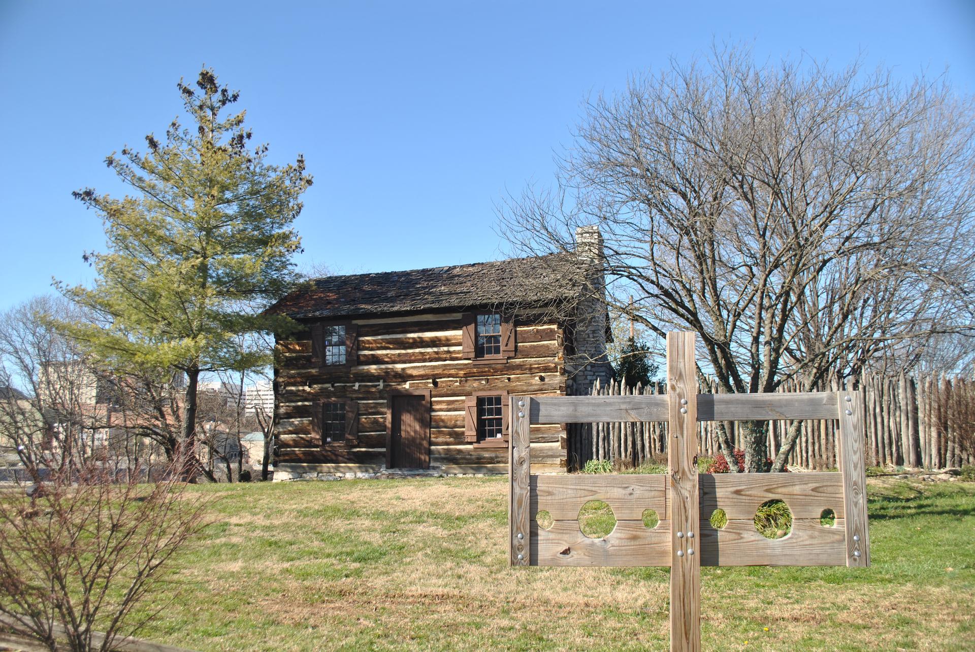 Historic log cabin next to wooden stocks in a sunny, grassy field.