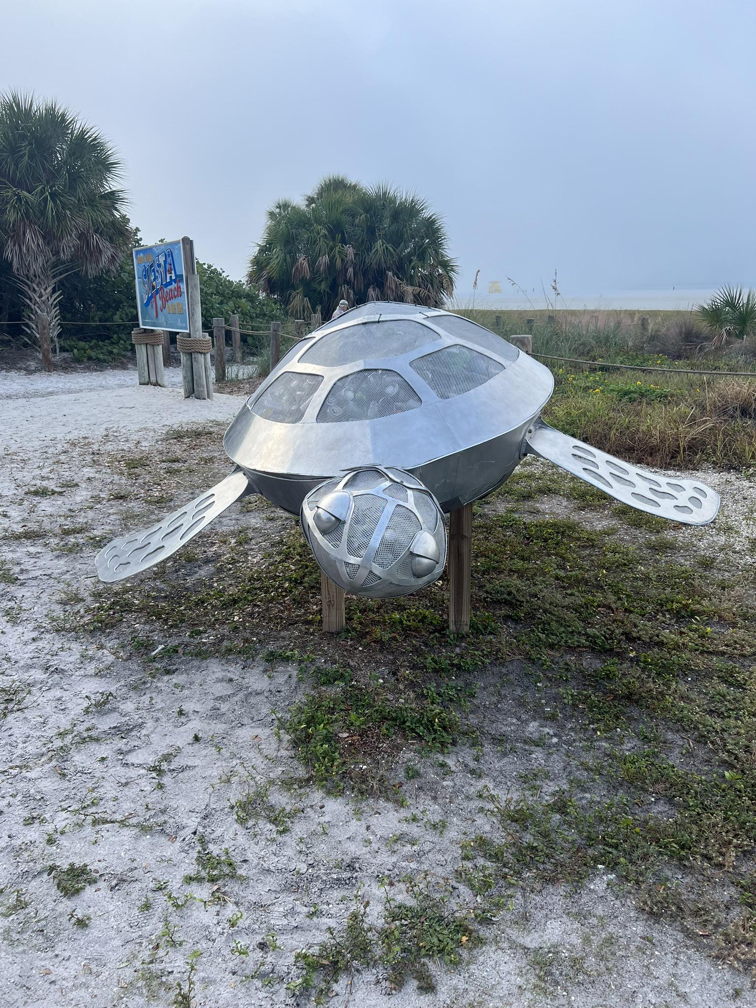 A large, shiny metal turtle sculpture on sandy ground near palm trees.