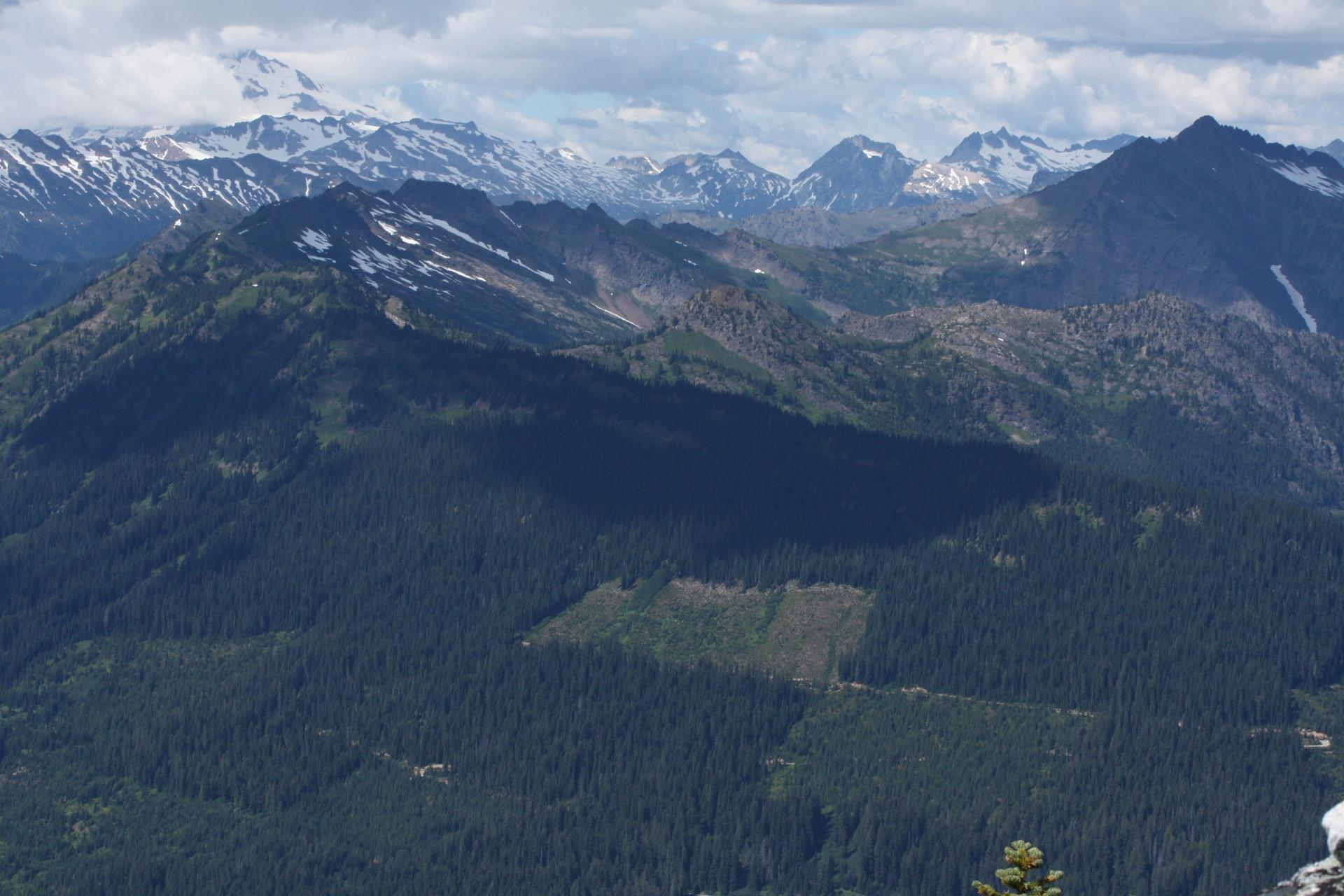Vast mountain landscape with dark forests, distant snowy peaks, and patchy sunlight.