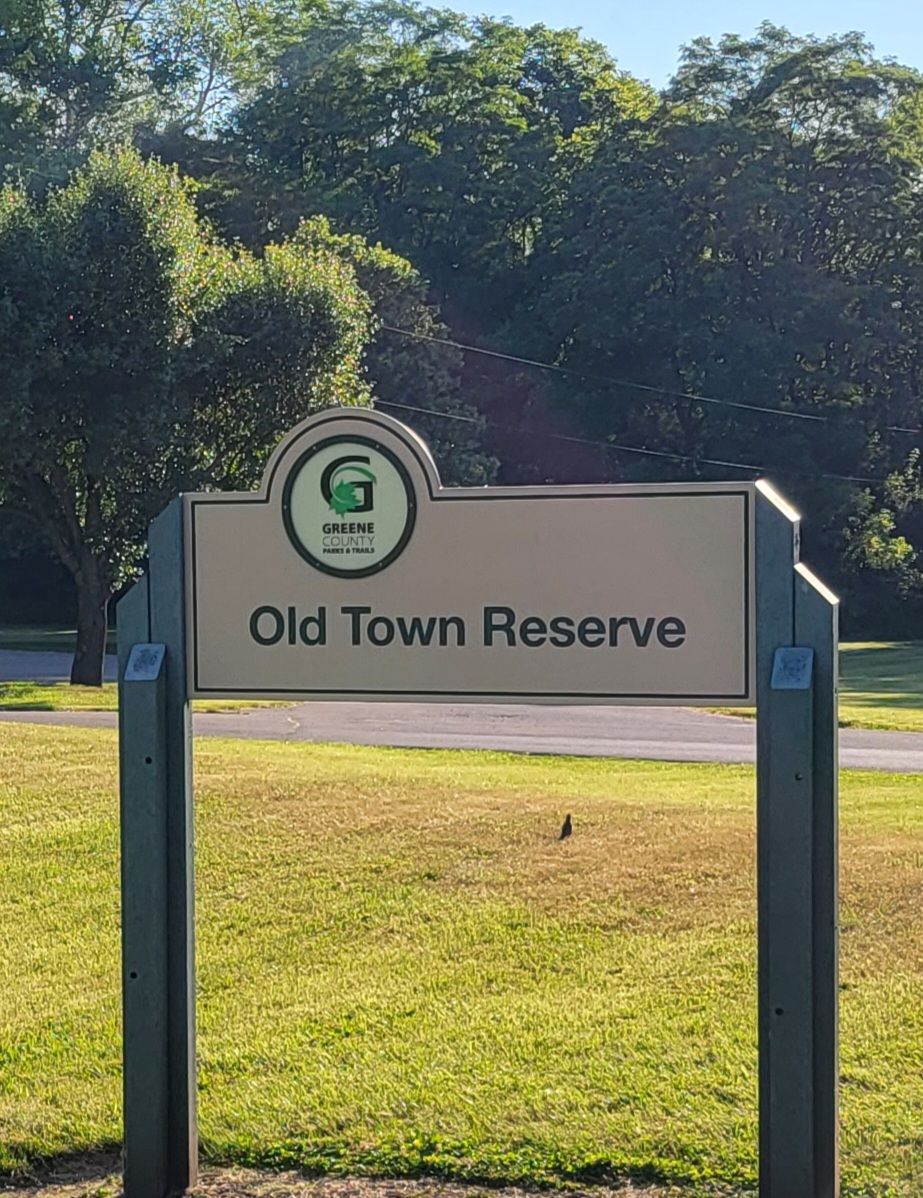A beige sign for a reserve stands in a sunny, grassy field with trees. - Little Miami Scenic Trail