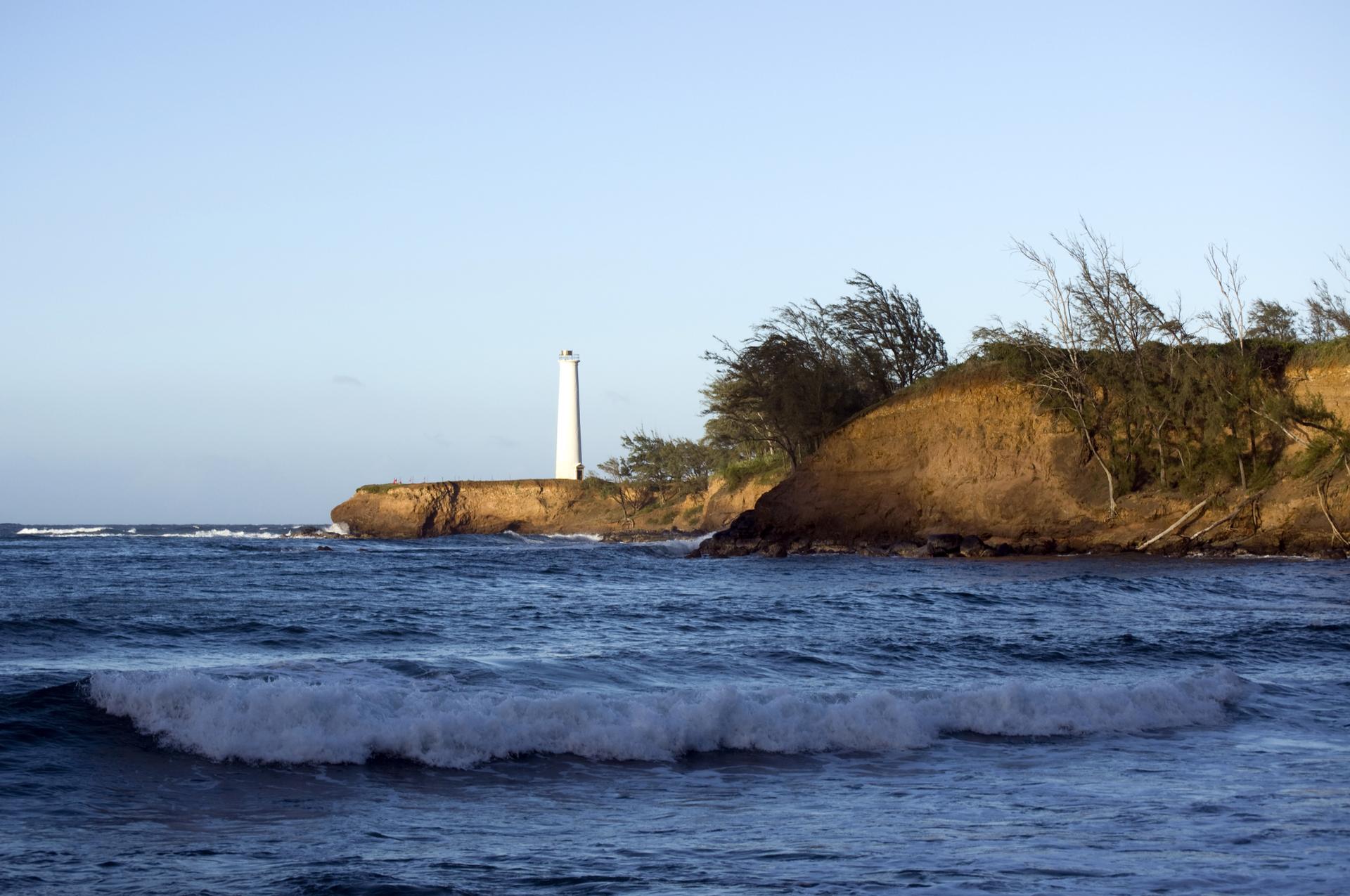 Ocean waves crash near a white lighthouse on a rocky, tree-lined cliff.